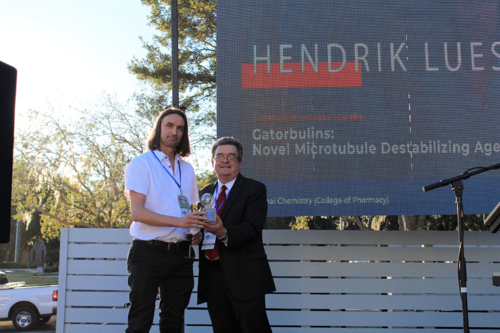 Research Hendrik Luesch stands with Dr. David Norton, VP of UF Research, after receiving the invention of the year award. They are on stage in front of a screen showing an image with Hendrick Luesch's name on it.