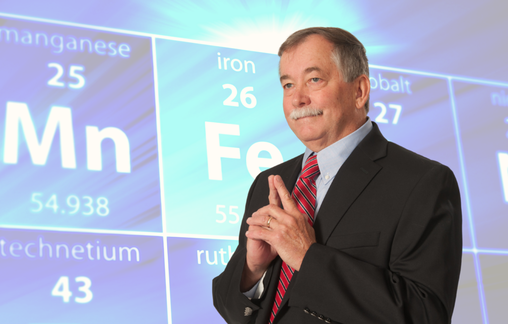 Dr. Raymond Bergeron stands with his two index fingers touching, as if in thought, in front of a giant periodic table with iron, right behind him