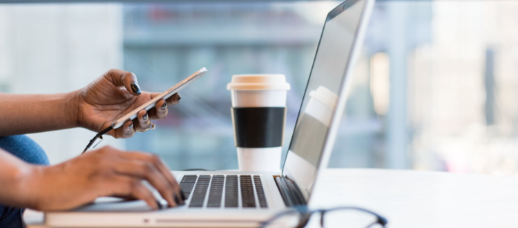 a woman of color -- seen only by her hands holding a phone and tapping on a laptop keyboard at a table with a pair of glasses and a coffee shop coffee cup beside her