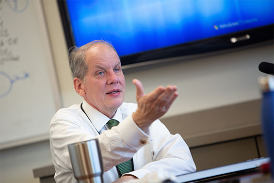 Dr. Mark Atkinson sits at a table, speaking and gesturing.
